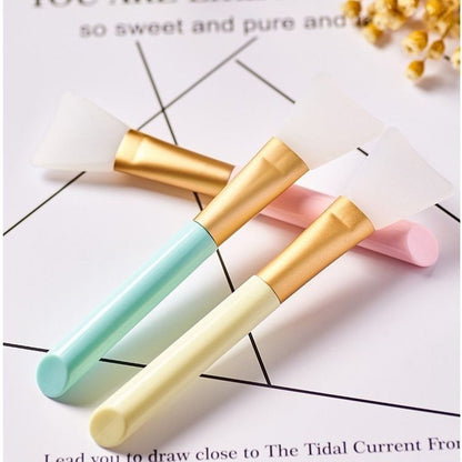 Three colorful facial silicone mask brushes with gold accents on a white background