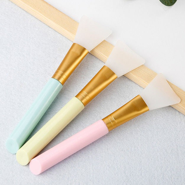 Three facial silicone mask brushes with pastel handles on a light gray background